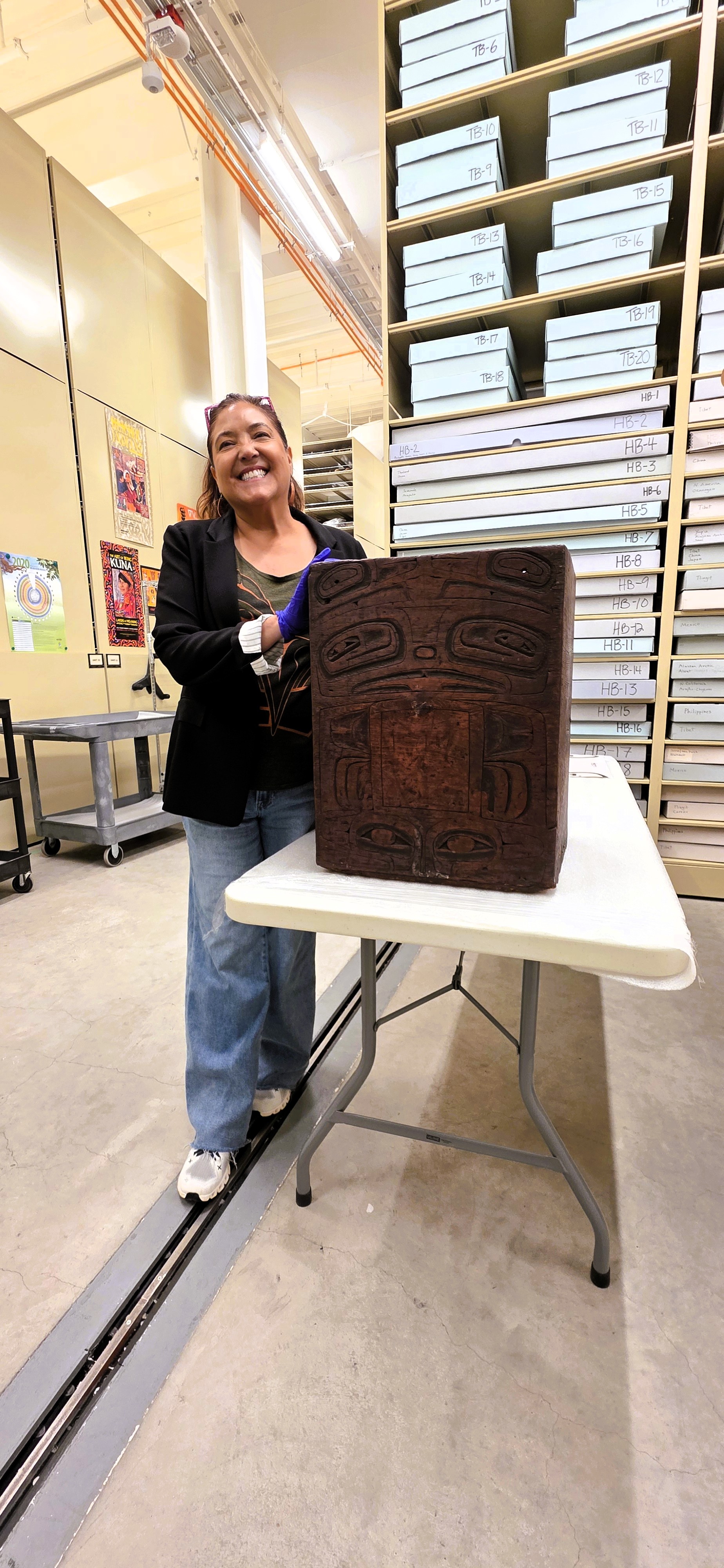 Tobi Iverson, wearing conservation gloves, standing beside a carved pre-contact bentwood box in the Burke Museum collections storage